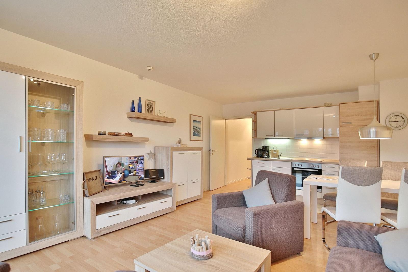 Wohnzimmer mit Küche und Esstisch. Fernseher, Schrank und Sessel sichtbar.
