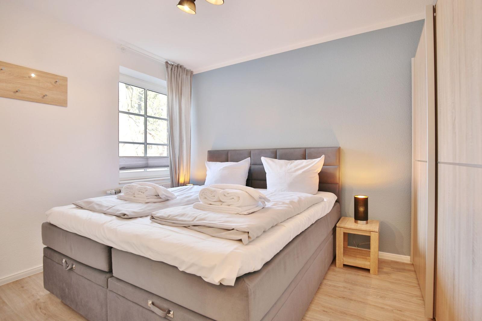Schlafzimmer mit Bett, Nachttisch und Fenster. Wandfarbe hellblau. Holzboden und Kleiderständer.