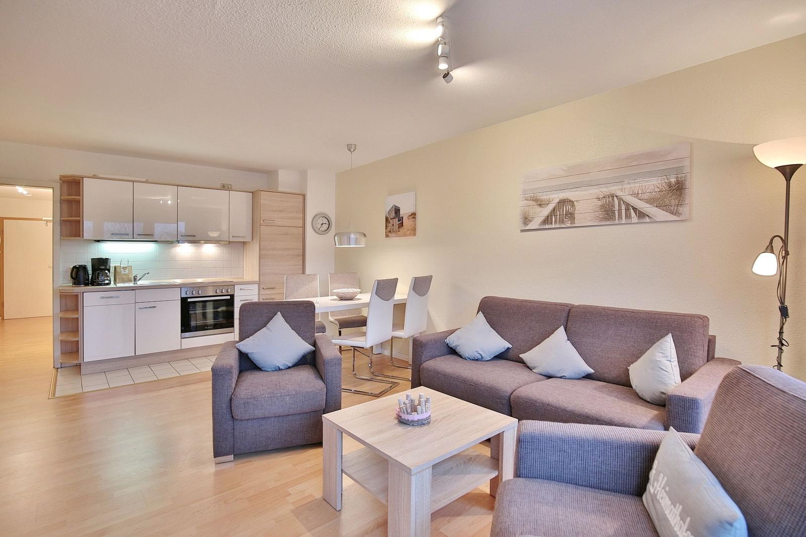 Wohnbereich mit Sofa, Sessel und Küchenbereich im Hintergrund.