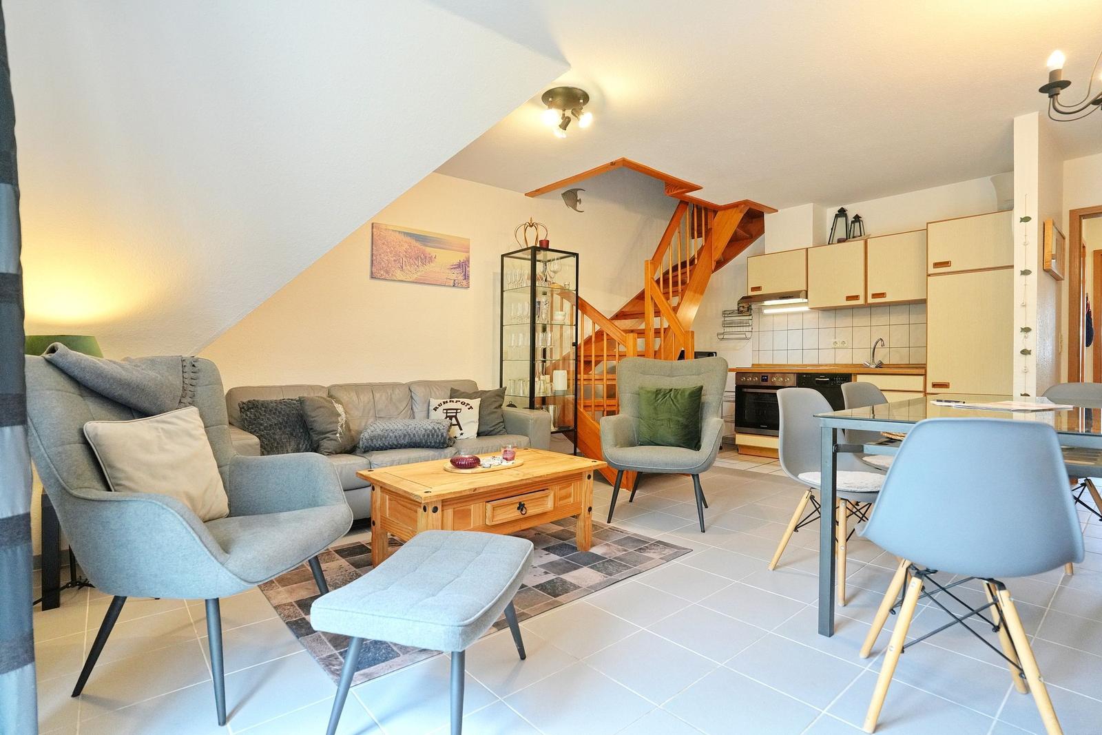 Wohnbereich mit Sofa, Sesseln und Essbereich. Holztreppe führt nach oben.