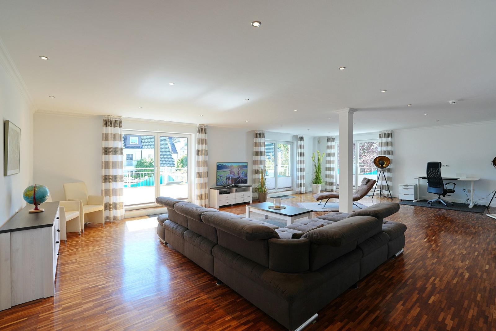 Großes Wohnzimmer mit Sofa, TV und Blick auf Balkon. Holzboden und großzügige Fenster.