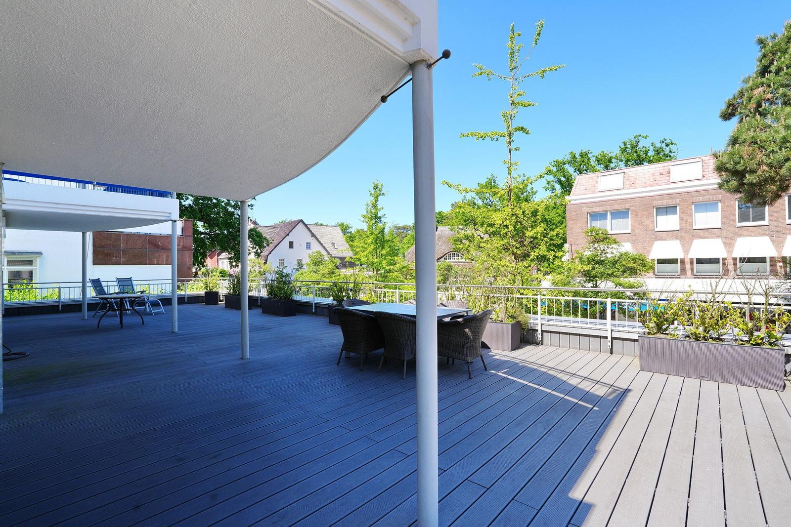 Terrasse mit Tisch, Stühlen und Sonnenschutz. Blick auf Grünflächen und Gebäude.