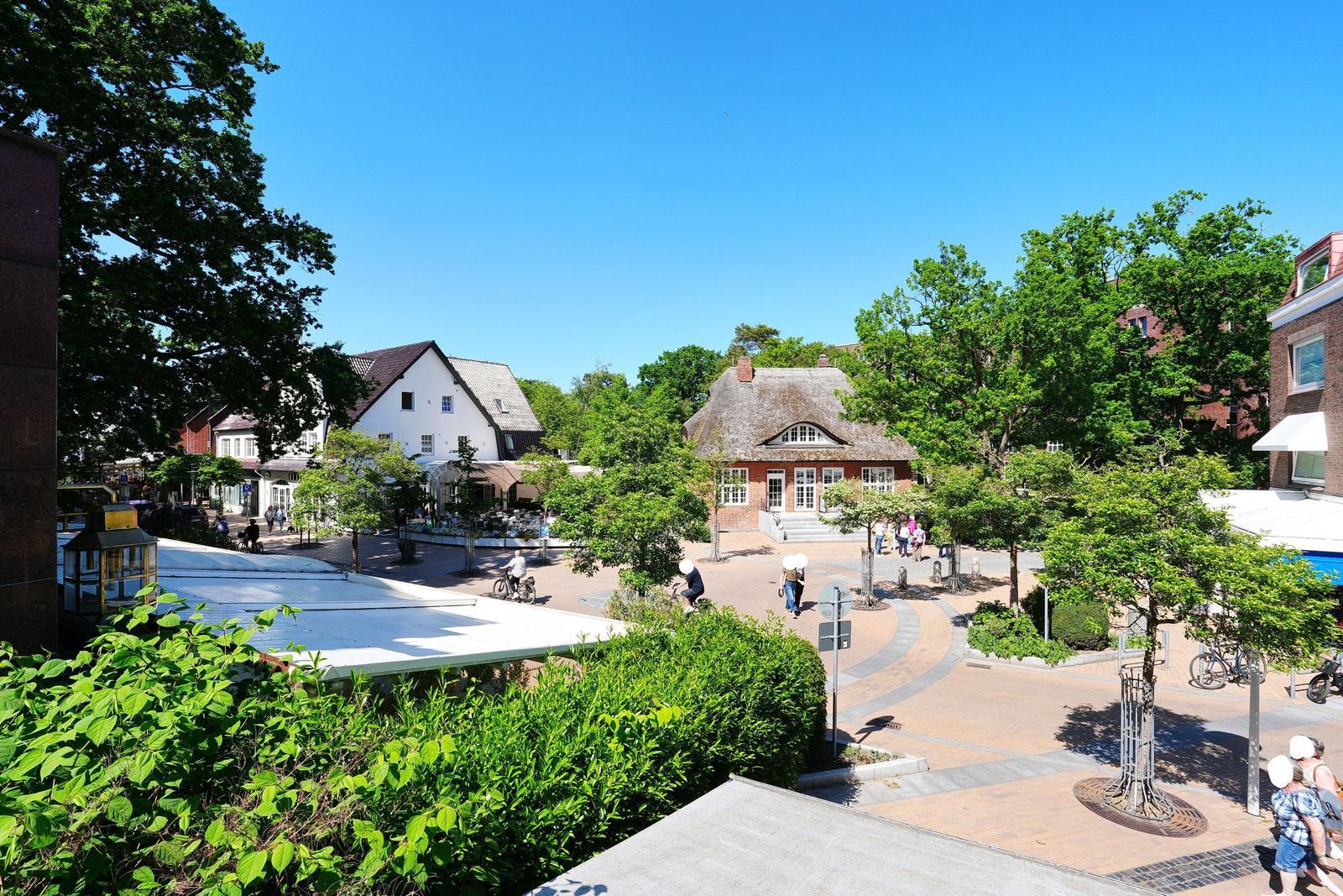 Stadtplatz mit Häusern, Bäumen und Spaziergängern unter blauem Himmel.