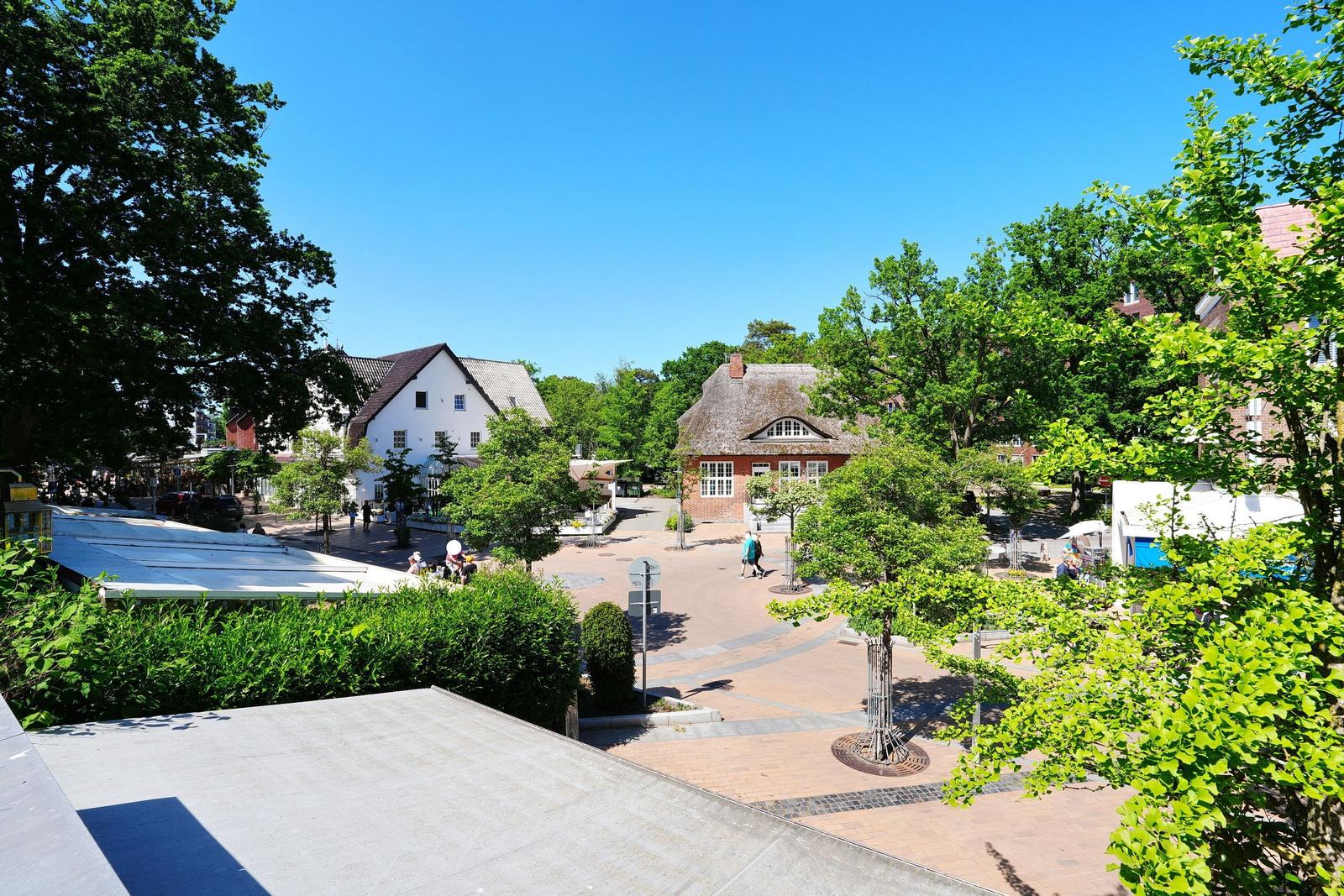 Platz mit Bäumen, Häusern und Menschen unter blauem Himmel.