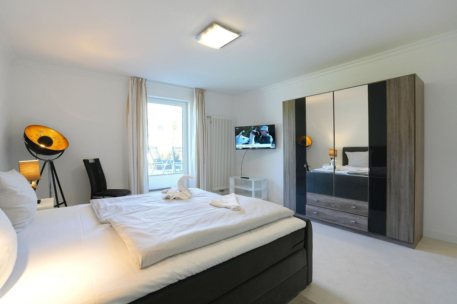 Schlafzimmer mit Doppelbett, TV, Kleiderschrank und Fenster mit Blick auf Balkon.