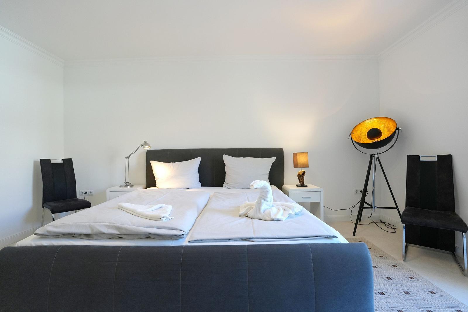 Schlafzimmer mit Doppelbett, zwei Nachttischen, Stehlampe und zwei Stühlen.