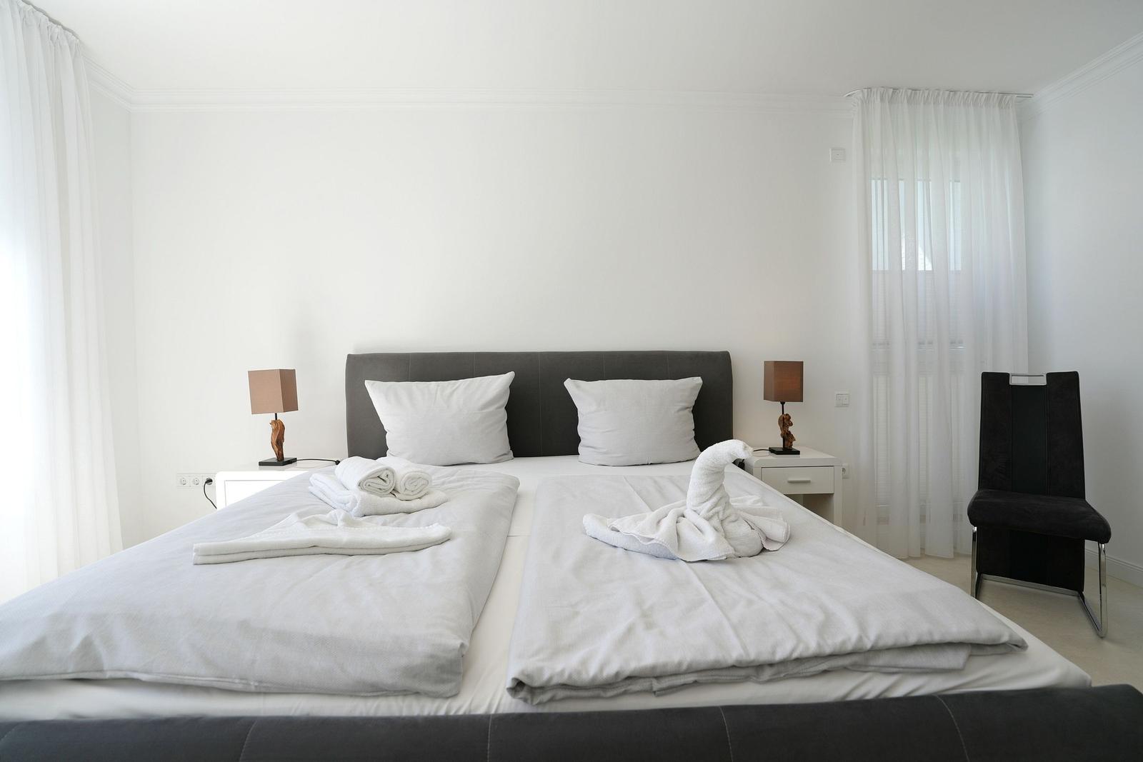 Schlafzimmer mit Doppelbett, zwei Nachttischen und Stuhl. Bett mit weißen Laken und Handtuchschwan.