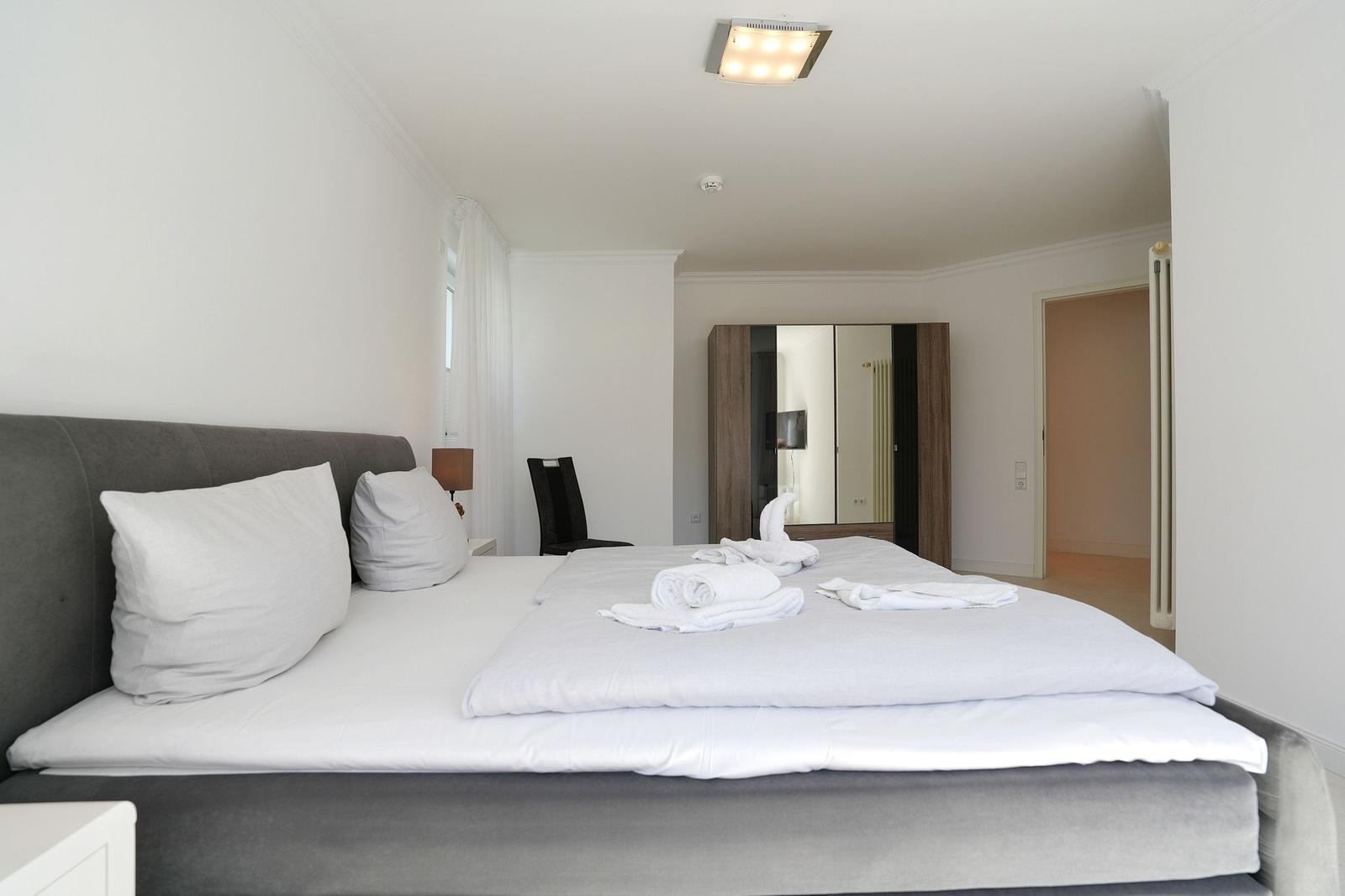 Schlafzimmer mit Bett, Kissen und Handtüchern. Spiegel und Stuhl sind sichtbar.