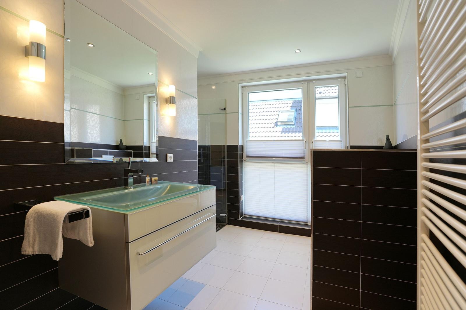 Badezimmer mit Glaswaschbecken, Spiegel und Dusche. Fenster mit Jalousie und Heizkörper.