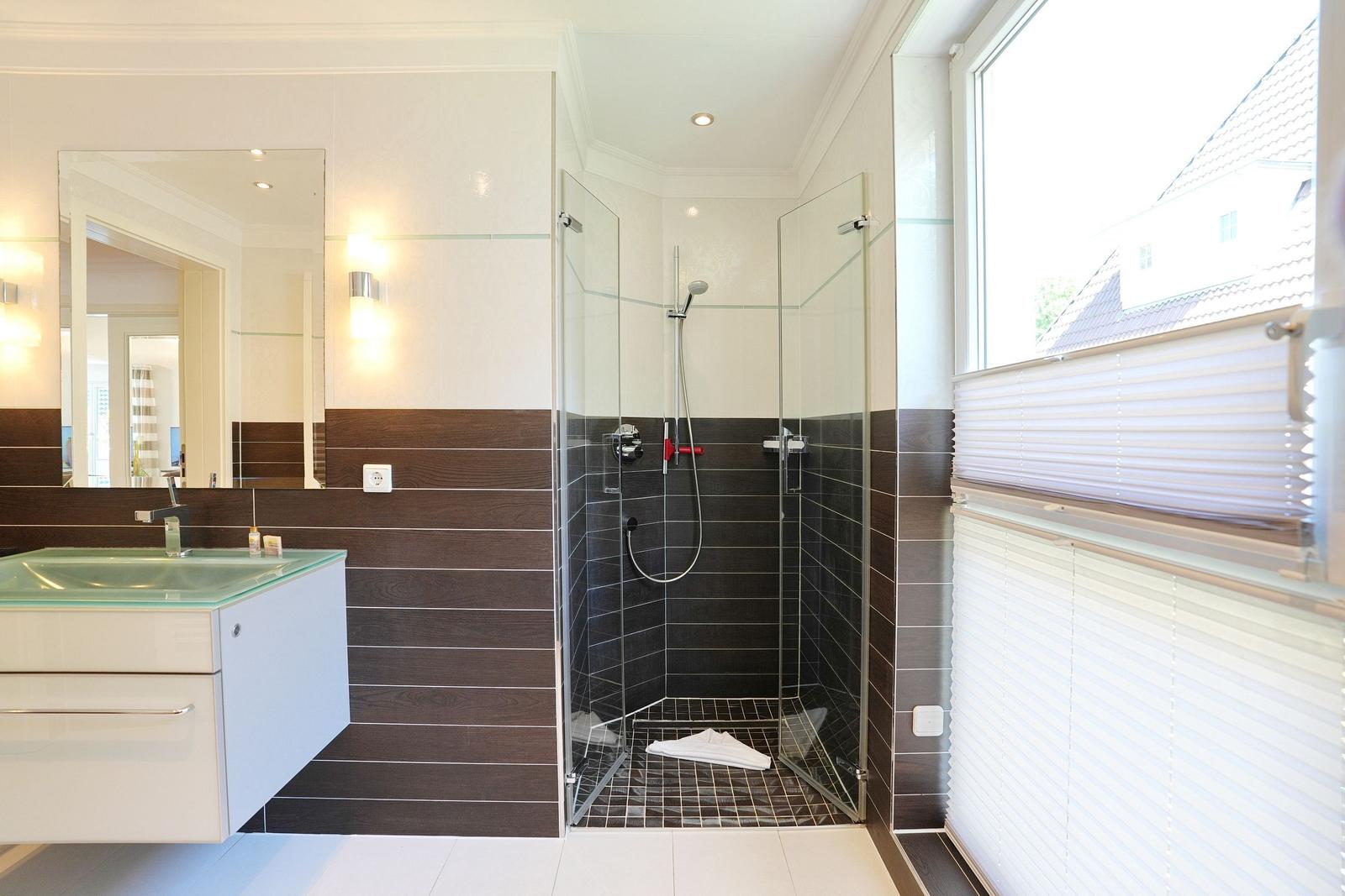 Badezimmer mit Dusche, Waschbecken und Fenster mit Jalousie.