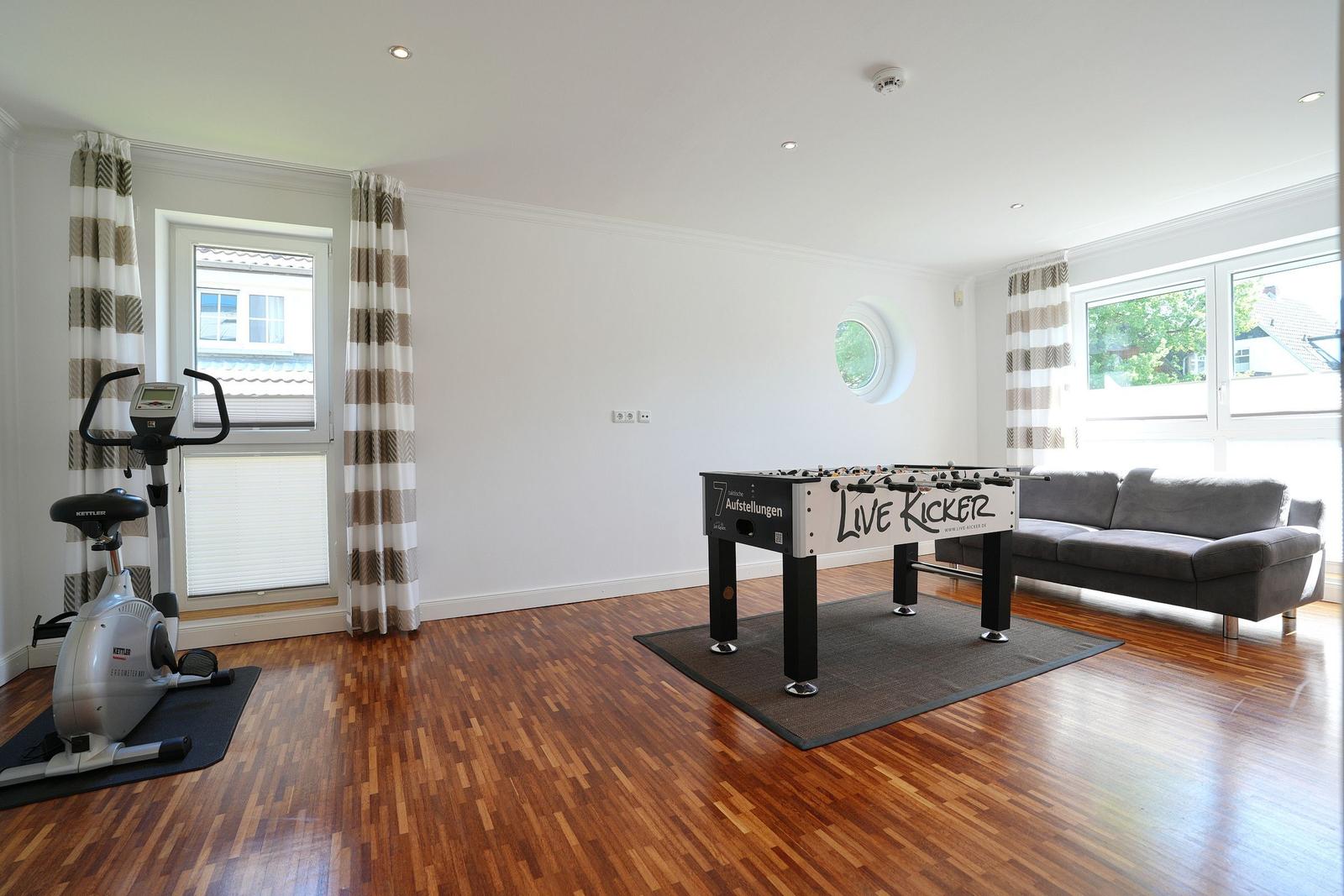 Raum mit Foosballtisch, Sofa, Fitnessrad und Holzboden. Fenster mit Vorhängen und runder Ausschnitt.