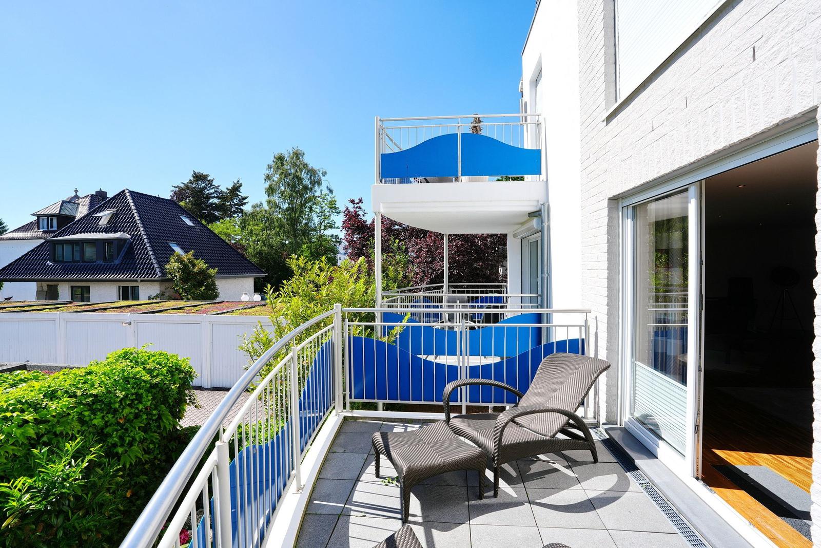 Terrasse mit Liegestühlen und blauem Sonnenschutz, Blick auf grüne Bäume und ein Haus.