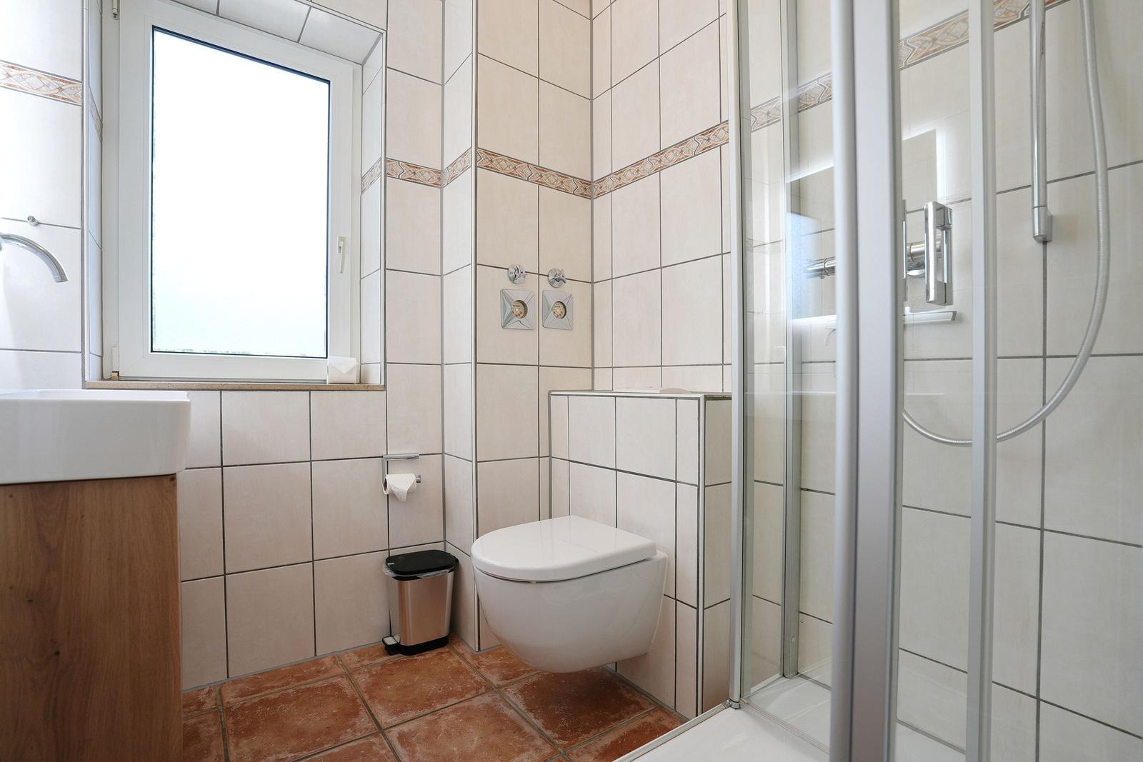 Badezimmer mit Dusche, Toilette und Waschbecken. Fliesen und Fenster sichtbar.