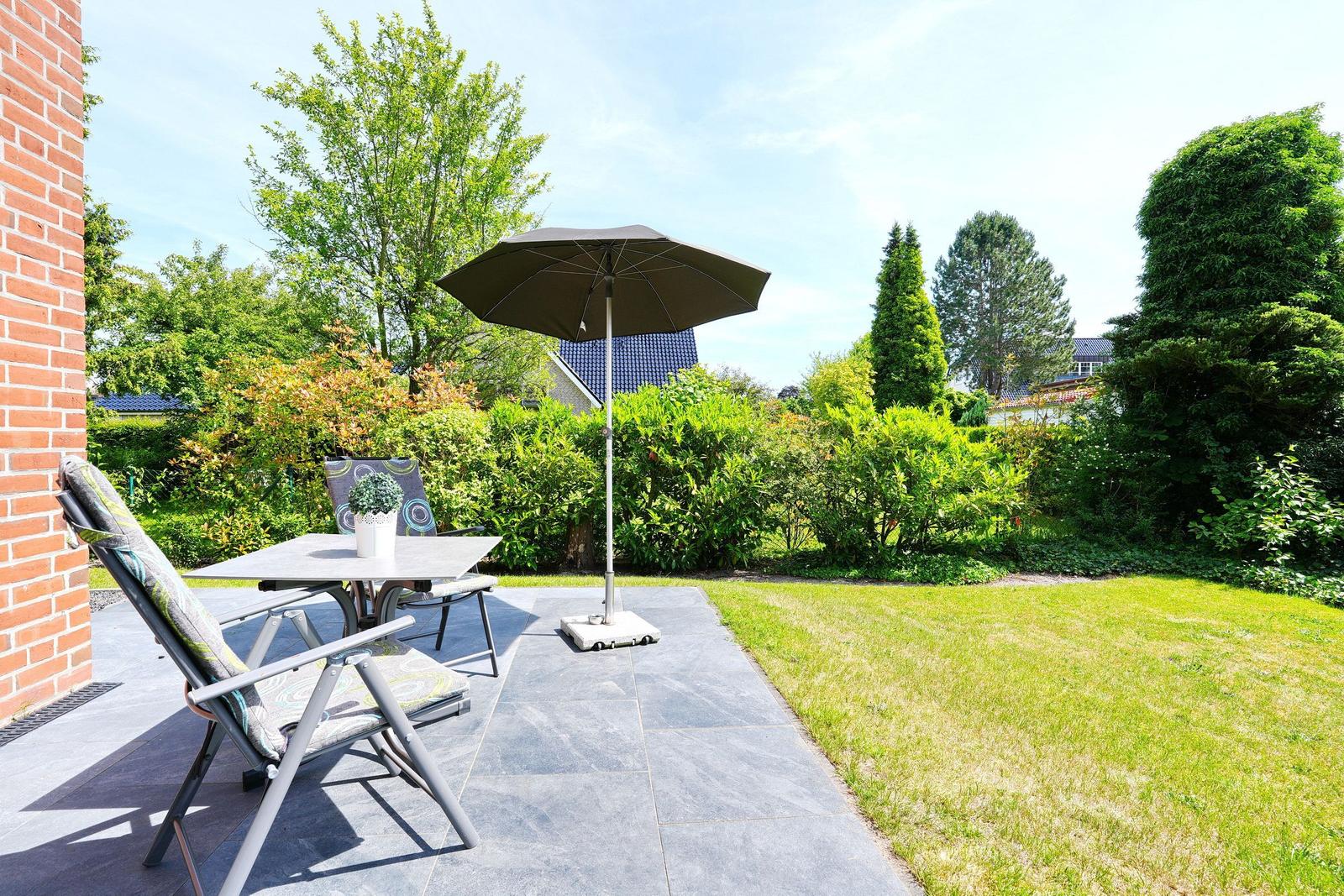 Terrasse mit Tisch, Stühlen und Sonnenschirm. Grünflächen und Bäume im Hintergrund.