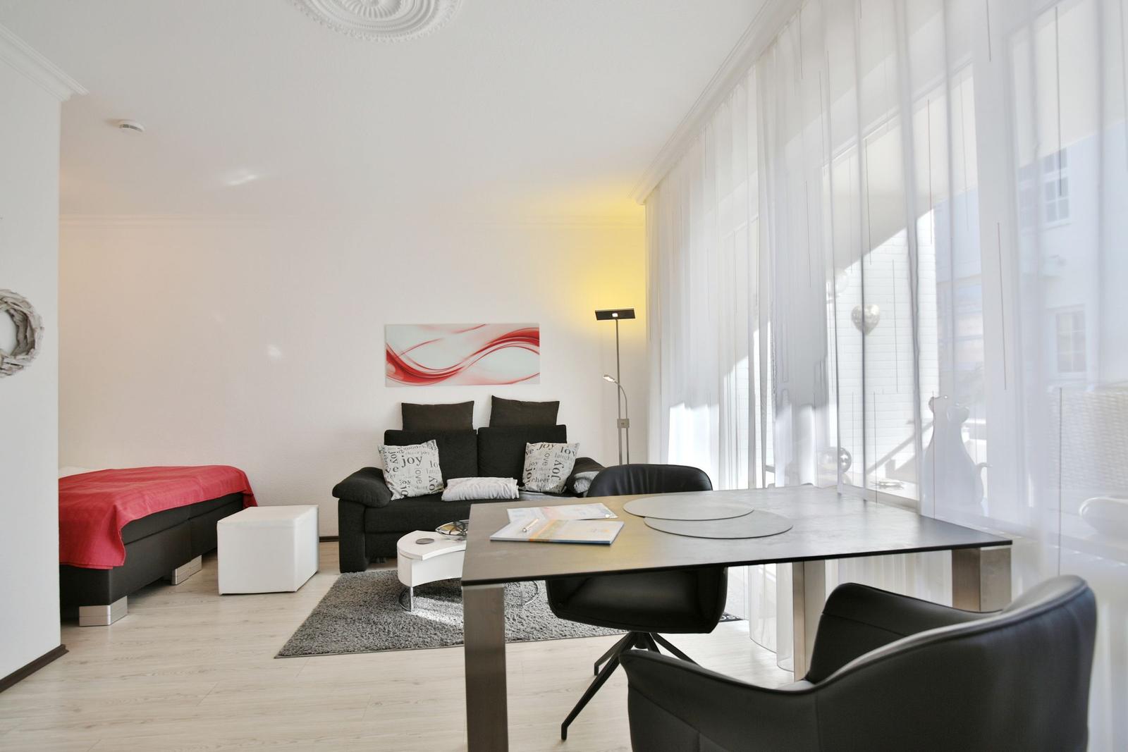Ein Raum mit Sofa, Tisch und Bett. Große Fenster mit weißen Vorhängen.