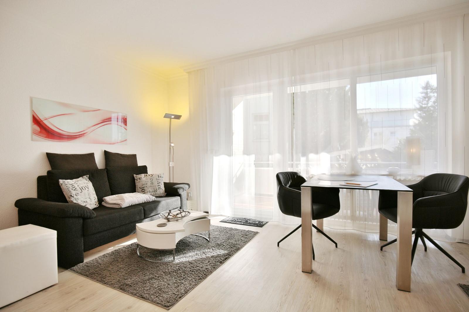 Wohnzimmer mit Sofa, Tisch und Stühlen. Fenster mit weißen Vorhängen.