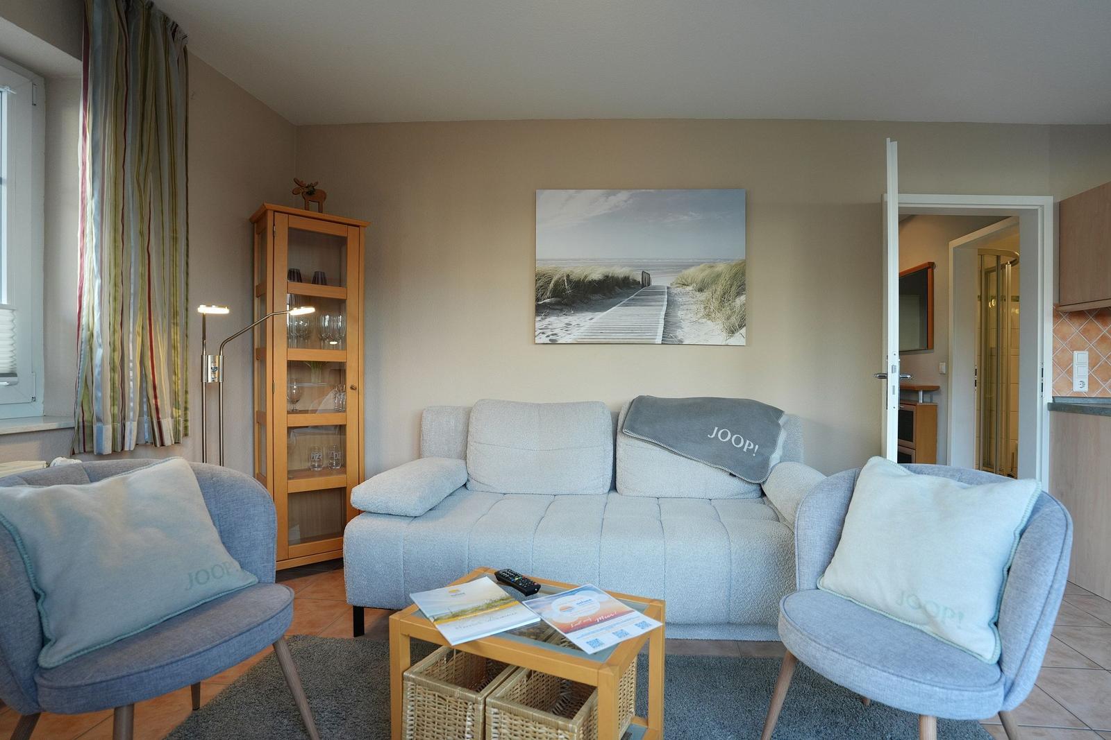 Wohnzimmer mit Sofa, zwei Sesseln und Glaswandregal. Wandbild zeigt Strand.