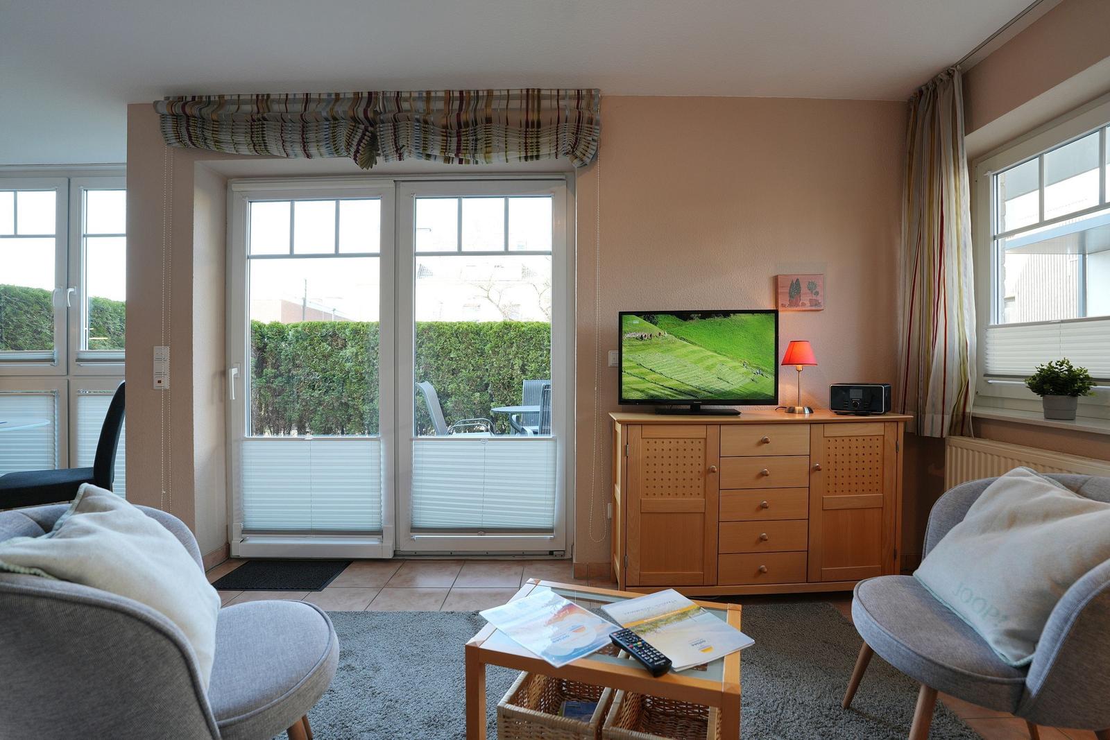 Wohnzimmer mit TV, zwei Sesseln und Blick auf Balkon mit Hecke.