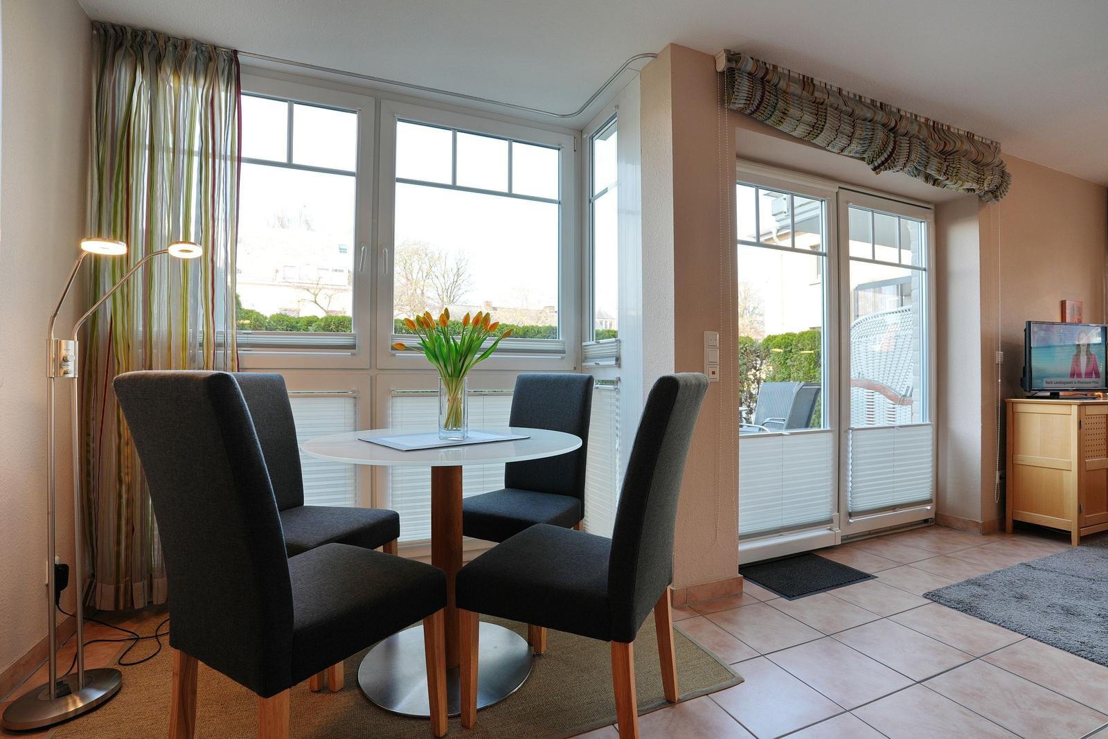Essbereich mit Tisch und Stühlen, Fenster und Balkontür. Blumen und Beleuchtung sichtbar.