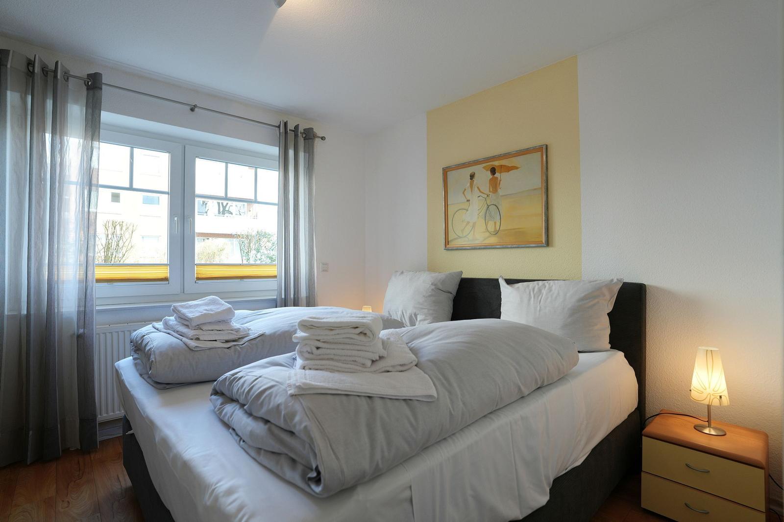Schlafzimmer mit Bett, Nachttisch und Fenster. Wandbild zeigt zwei Personen mit Fahrrad.