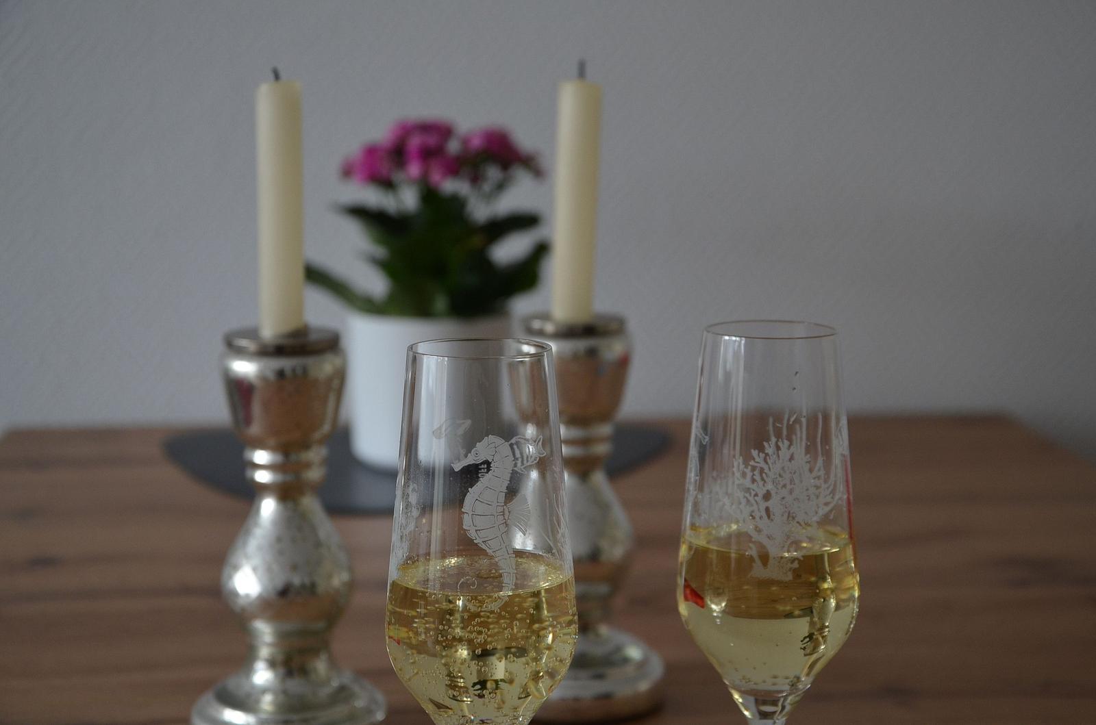 Zwei Gläser mit Sekt stehen auf einem Tisch. Im Hintergrund sind Kerzen und eine Pflanze zu sehen.
