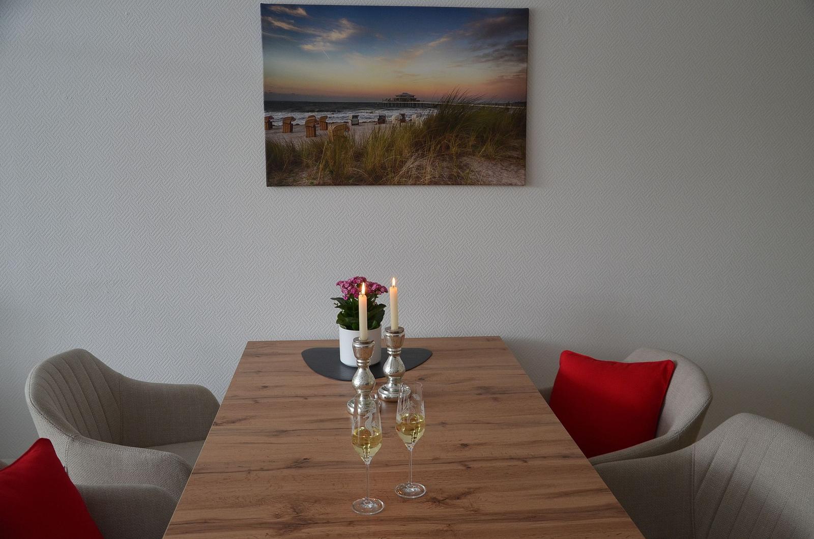 Esstisch mit zwei Stühlen, roten Kissen und Kerzen. Wandbild zeigt Strand und Pier.