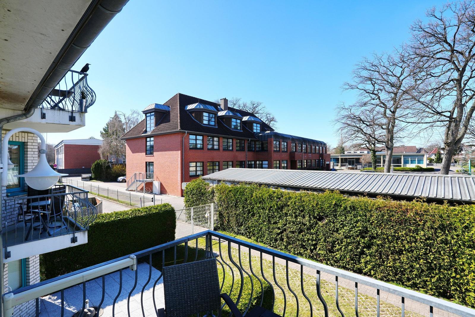 Balkon mit Gartenblick auf roten Backsteinbau mit Dachgauben und grünen Hecken.