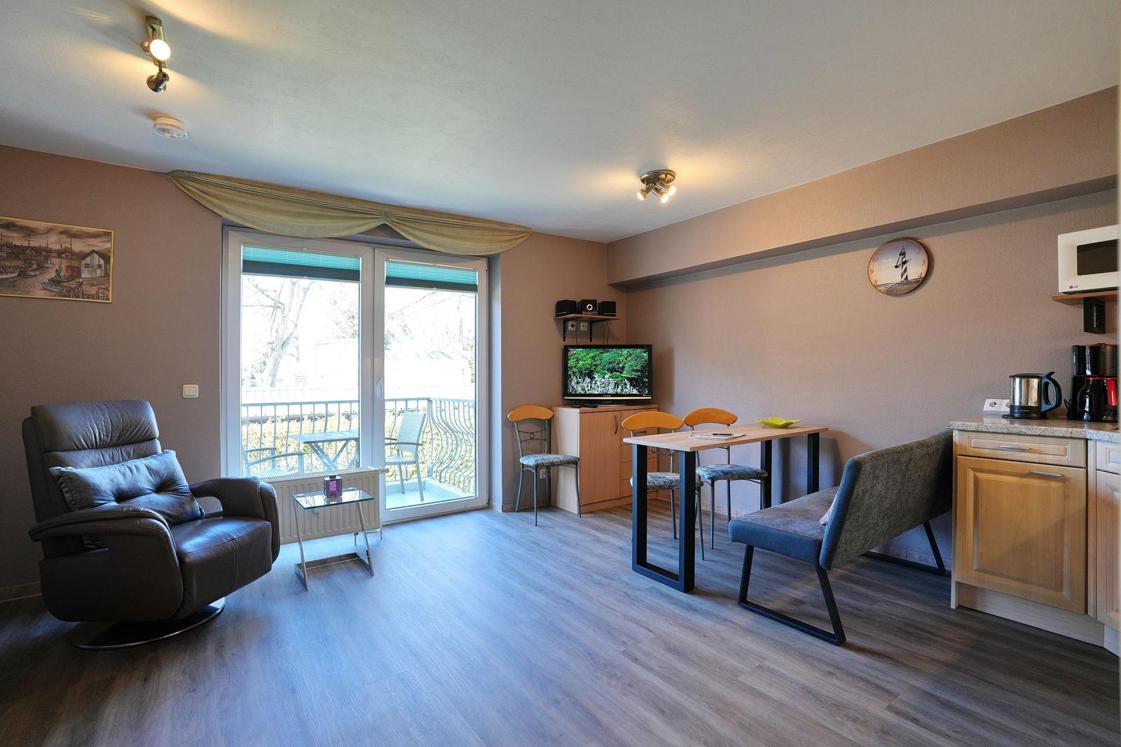 Wohnzimmer mit Essbereich, Küche und Balkon. Fernseher, Stühle und Sofa sind vorhanden.
