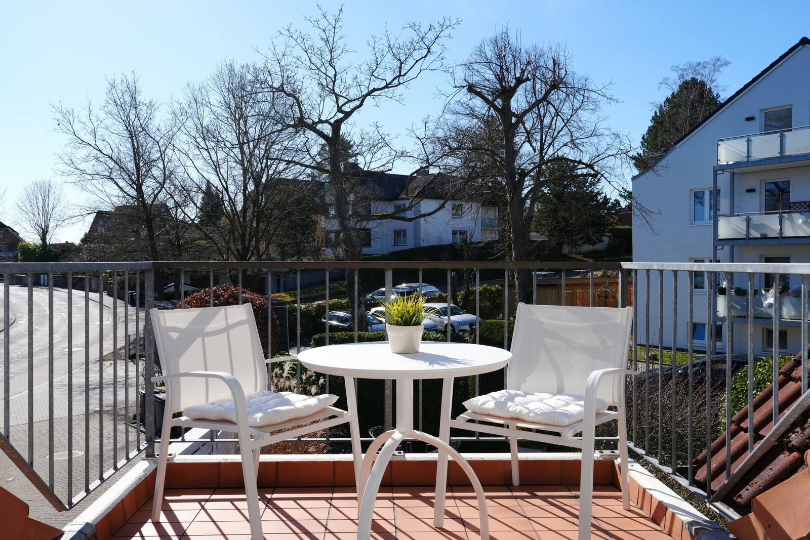 Terrasse mit weißem Tisch und zwei Stühlen, Blick auf Straße und Bäume.