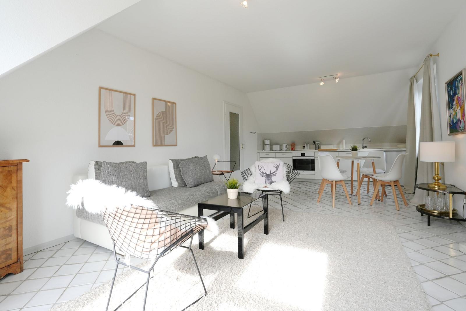 Wohnung mit Wohn- und Küchenbereich, Sofa, Stühlen und Tisch. Weiße Wände und Bodenfliesen.