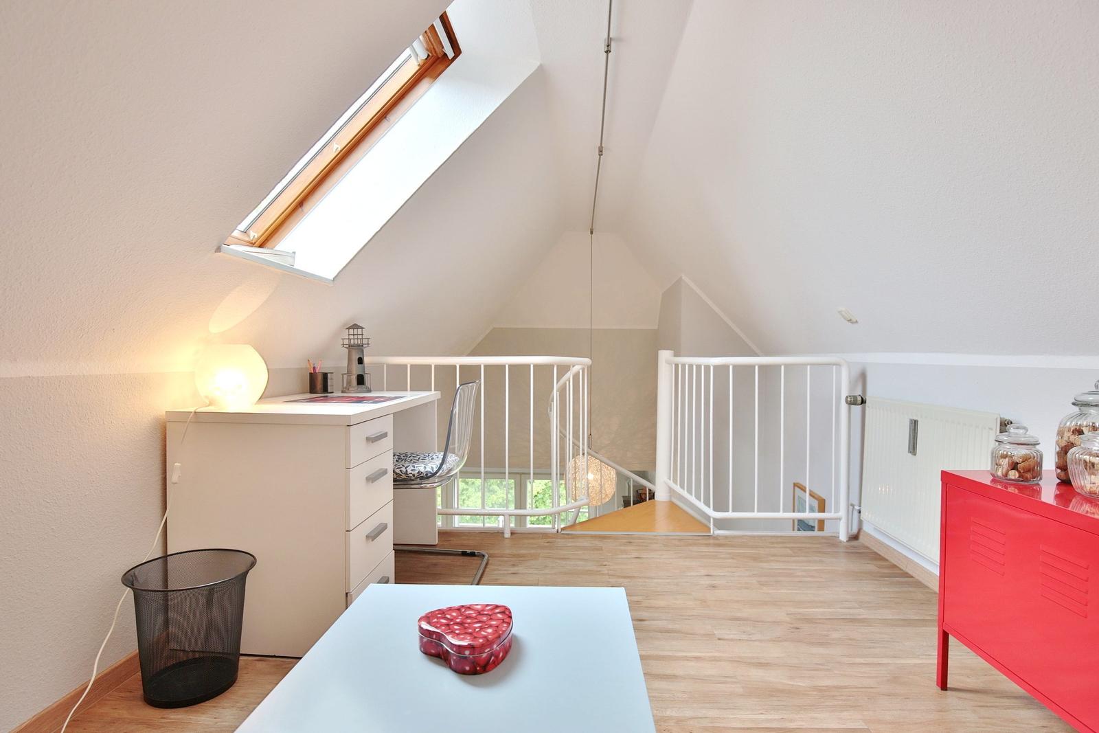 A bedroom with sloped ceilings, desk, red cabinet, and window.