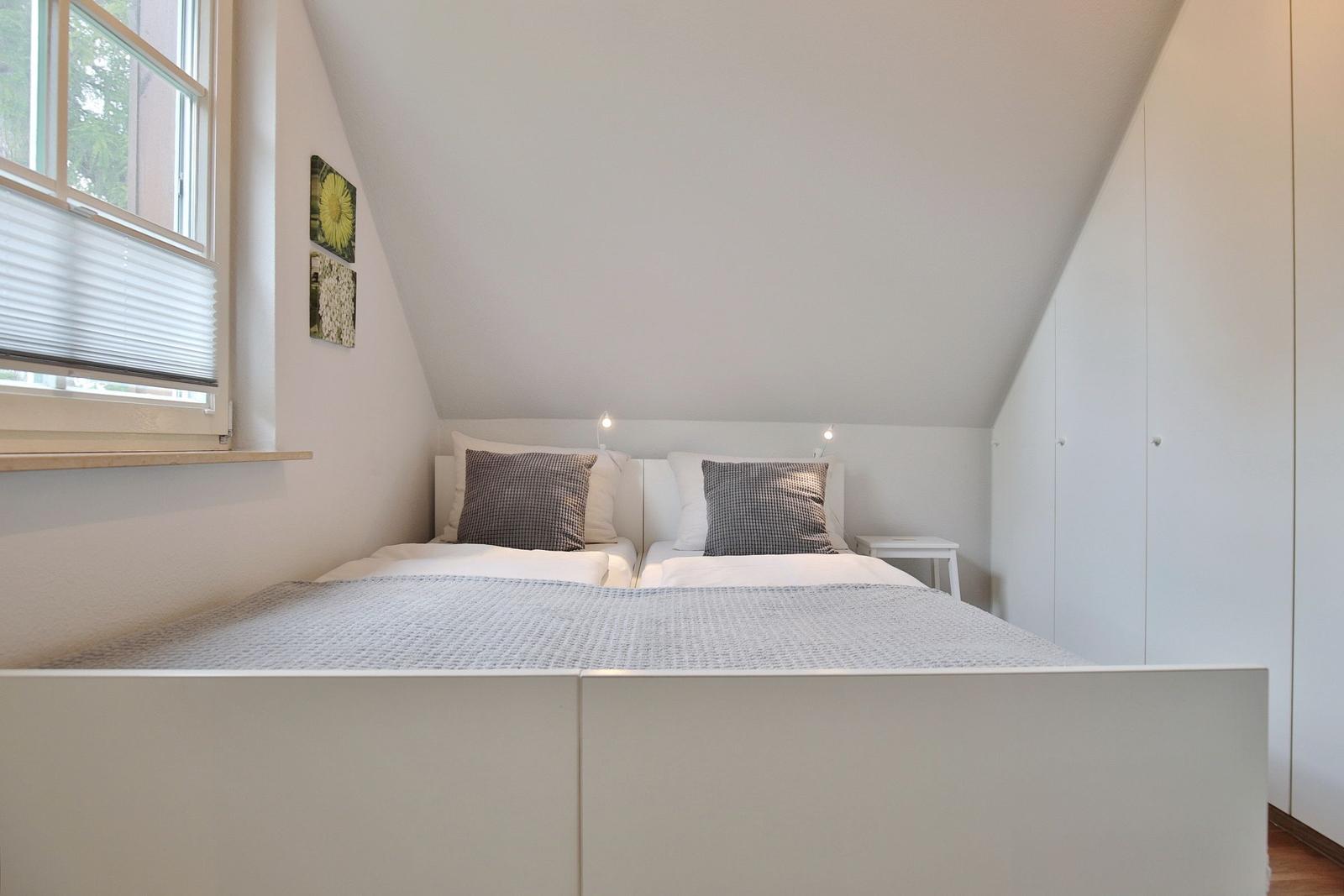 Bedroom with sloped ceiling, white bed, and window with blinds.