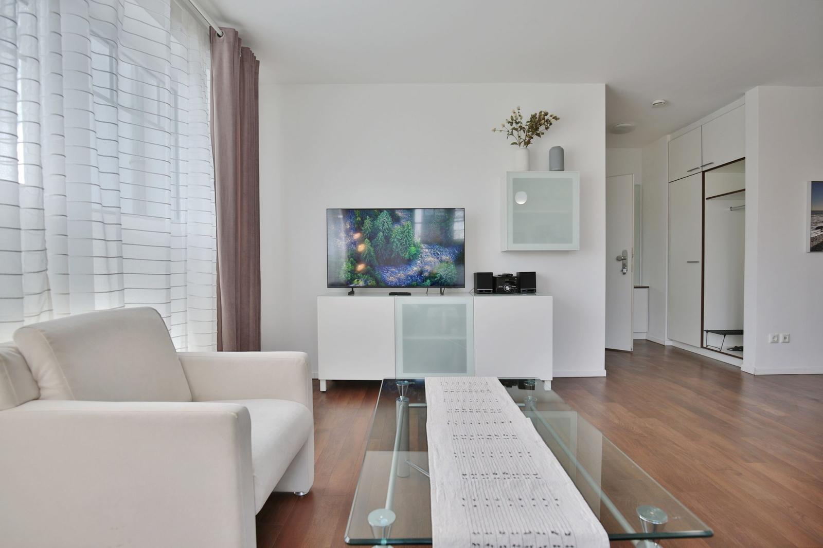 Wohnzimmer mit Sofa, Fernseher und Glas-Tisch. Holzfußboden und weiße Wände.