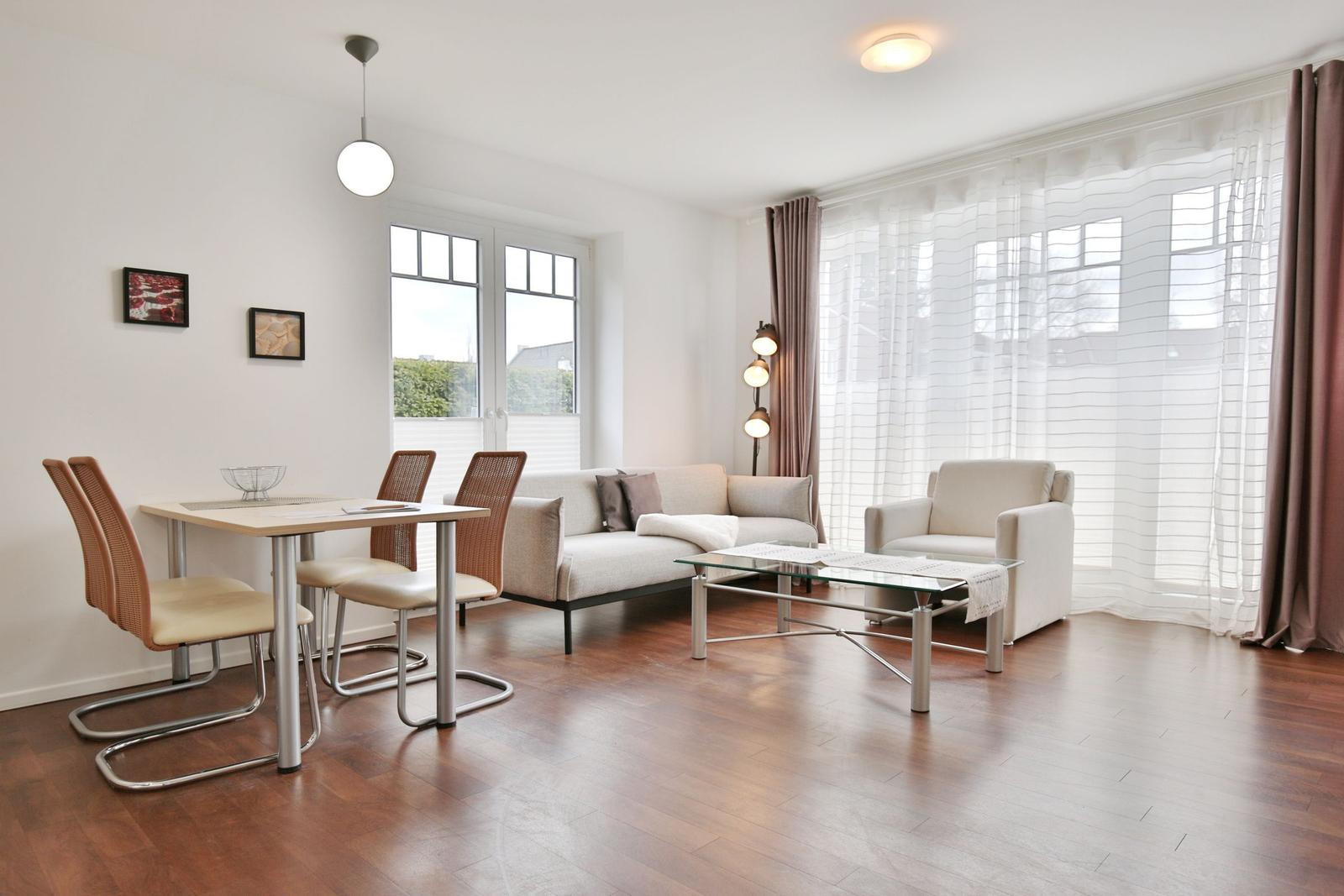 Wohn- und Essbereich mit Sofa, Sessel, Tisch und Stühlen. Holzboden und große Fenster.