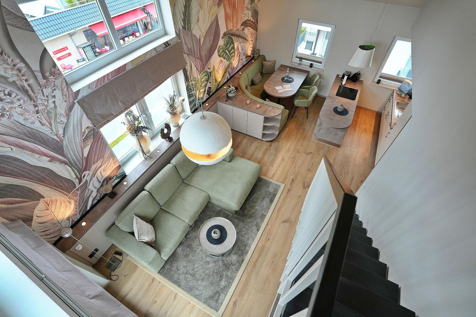 Living area with sofa, dining table, and kitchen. Wallpaper with plant motif. Wooden floor and large windows.