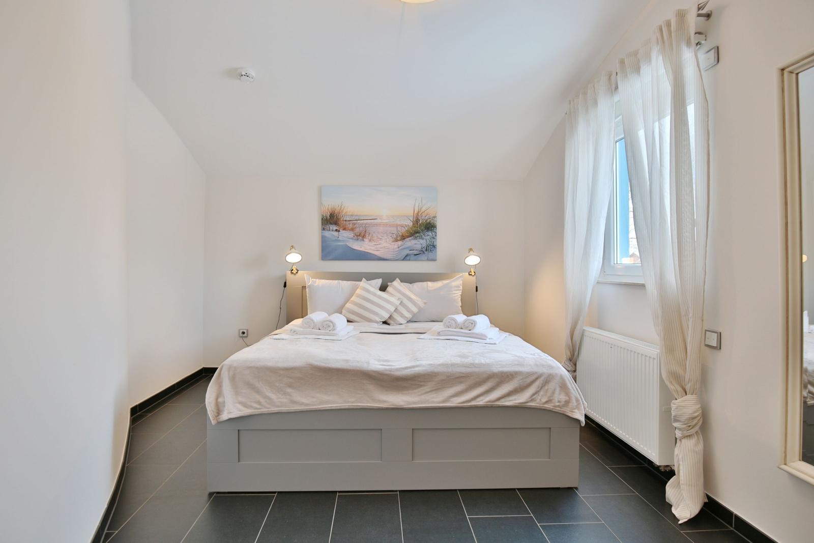 Schlafzimmer mit Doppelbett, Fenster und Wandbild. Fliesenboden und weiße Wände.