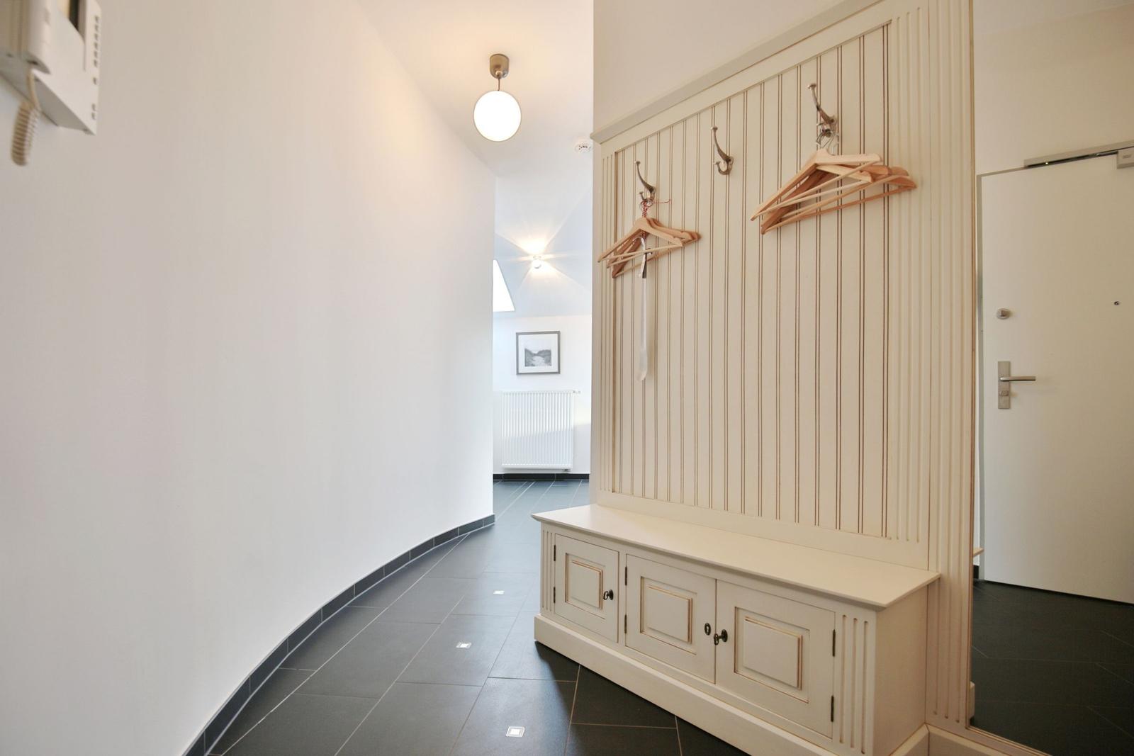 Eingangsbereich mit Garderobe, Haken und Sitzbank. Fliesenboden und weiße Wände.