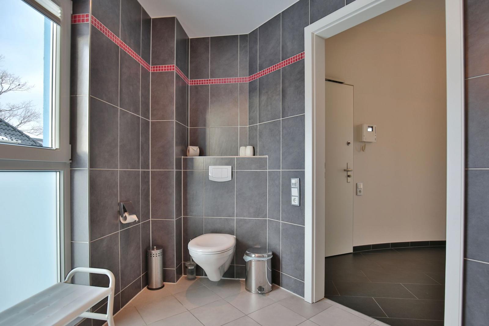 Badezimmer mit Toilette, Fenster und grauen Fliesen mit roter Zierleiste.