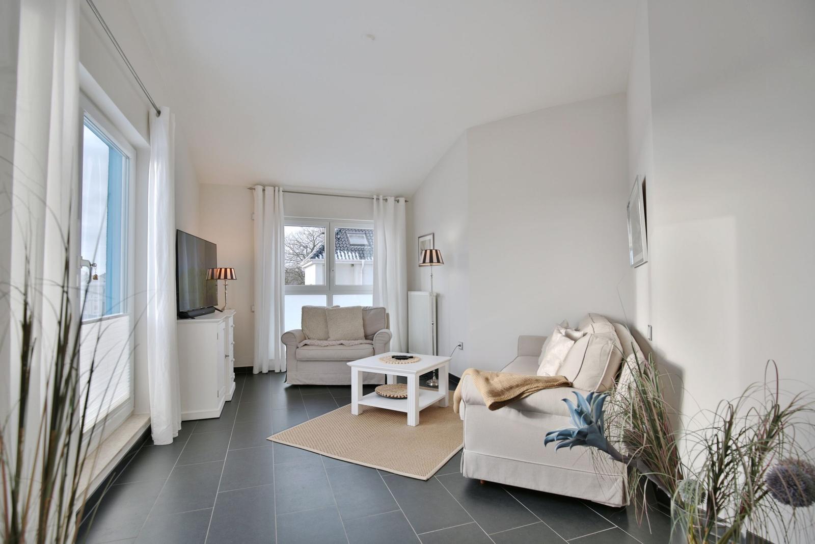 Wohnzimmer mit Sofa, Sessel und TV. Fenster mit weißen Vorhängen. Teppich und Pflanzen.
