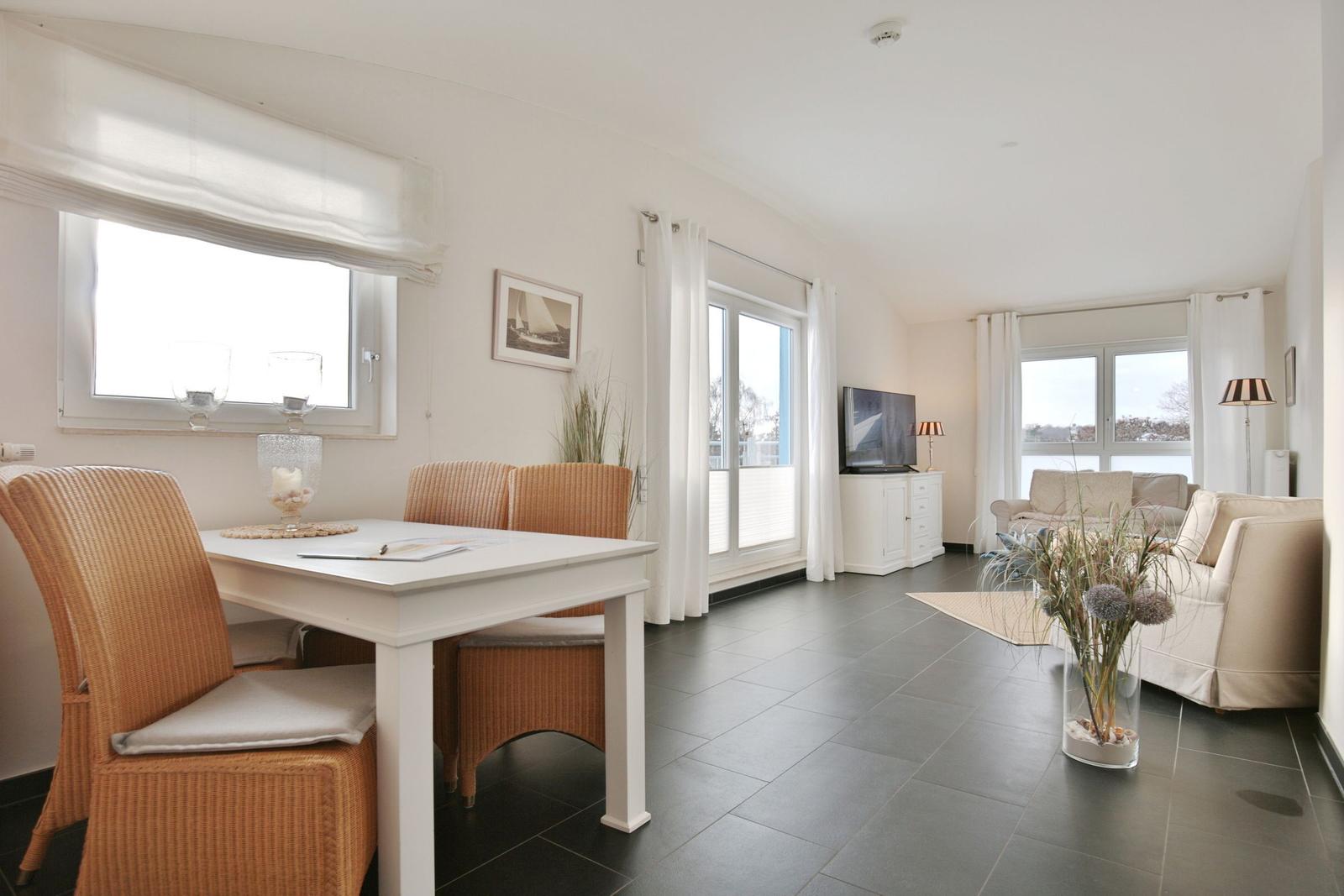 Wohn- und Essbereich mit Tisch, Stühlen und Sofa. Fenster und Glastüren mit weißen Vorhängen.