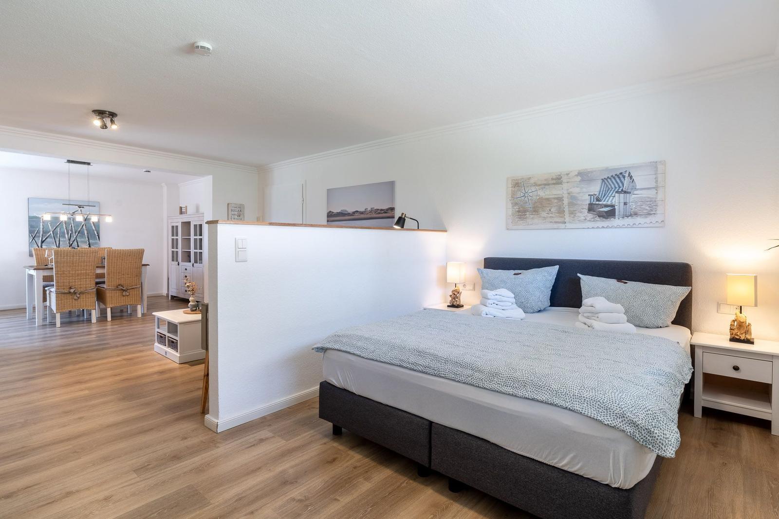 Schlafzimmer mit Doppelbett, Nachttischen und Wandbildern. Durchgang zur Küche mit Esstisch.