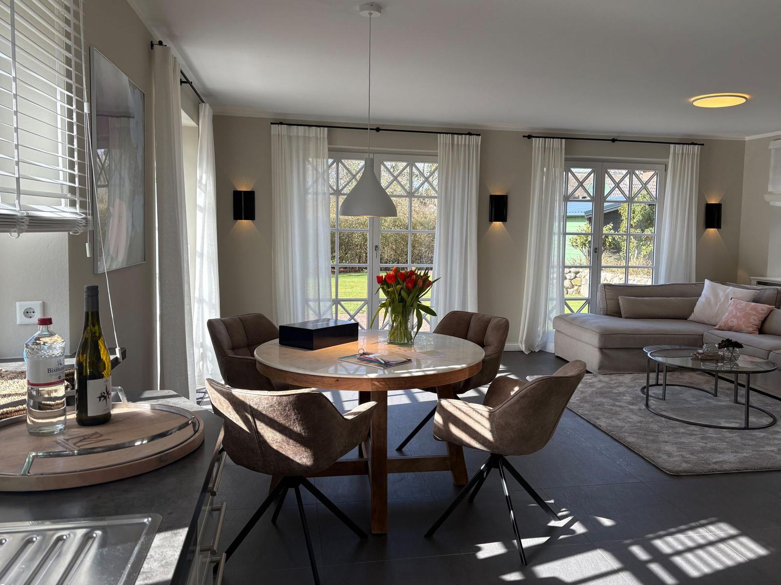Essbereich mit runden Tisch und vier Stühlen. Wohnbereich mit Sofa und Glas-Tisch. Fenster mit weißen Vorhängen.