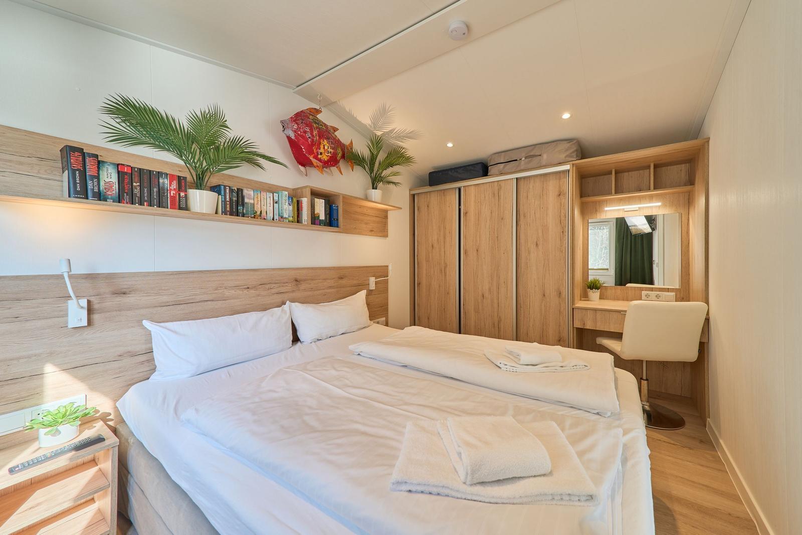 Schlafzimmer mit Bett, Kleiderschrank und Schreibtisch. Bücherregal und Pflanzen an der Wand.
