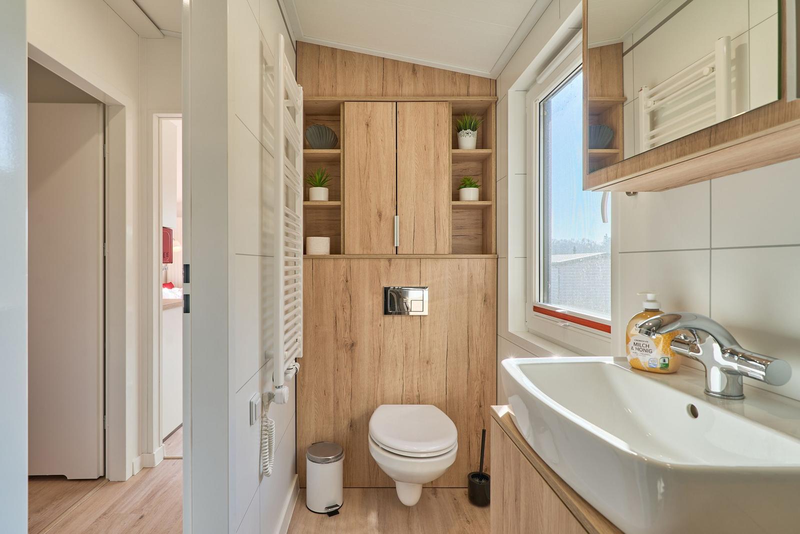 Badezimmer mit Toilette, Waschbecken und Holzregalen. Fenster mit Blick nach draußen.