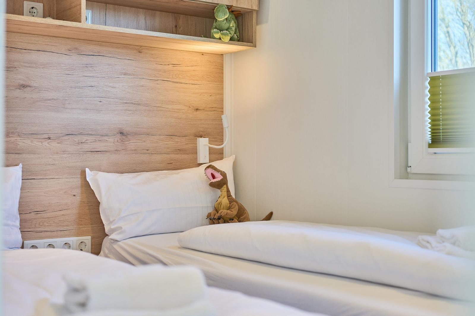 Schlafzimmer mit Holzwand, Bett und Fenster mit Jalousie. Auf dem Bett sitzt ein Stofftier.