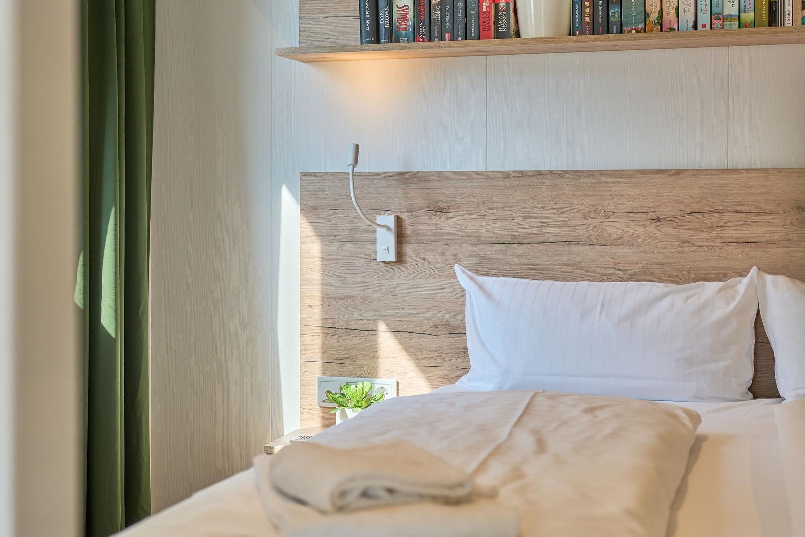 Schlafzimmer mit Holzkopfteil, Bett mit weißen Kissen und Decke, Wandregal mit Büchern und grüne Vorhänge.