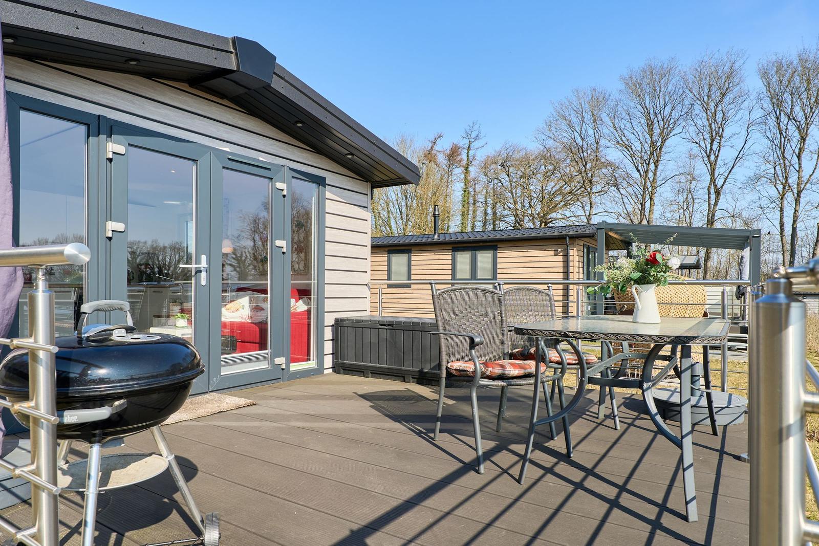 Terrasse mit Grill, Tisch und Stühlen. Hintergrund: Holzhaus und Bäume.