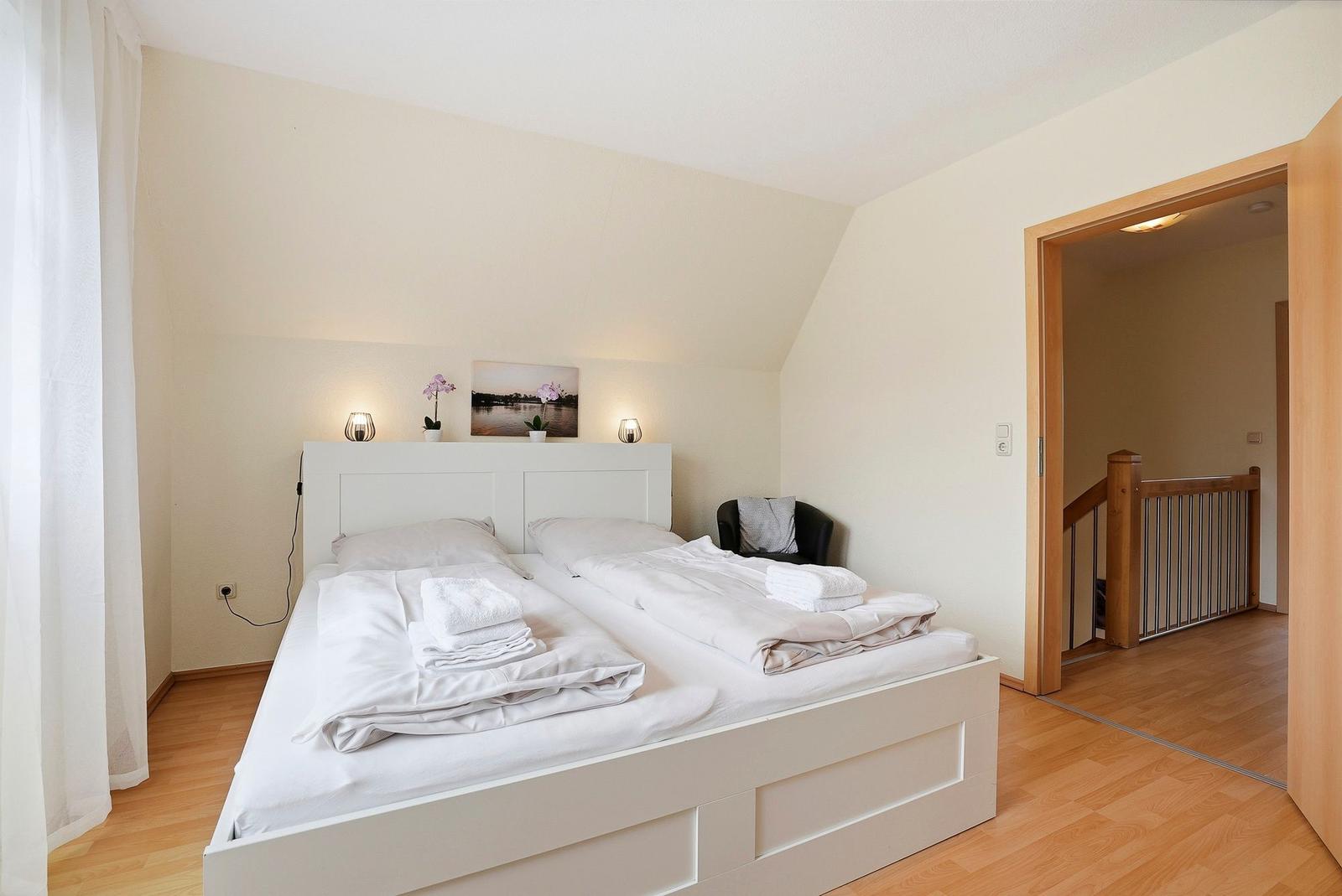 Schlafzimmer mit Doppelbett, weißen Bettwäsche und Holzboden. Tür führt in einen Flur mit Treppenhaus.
