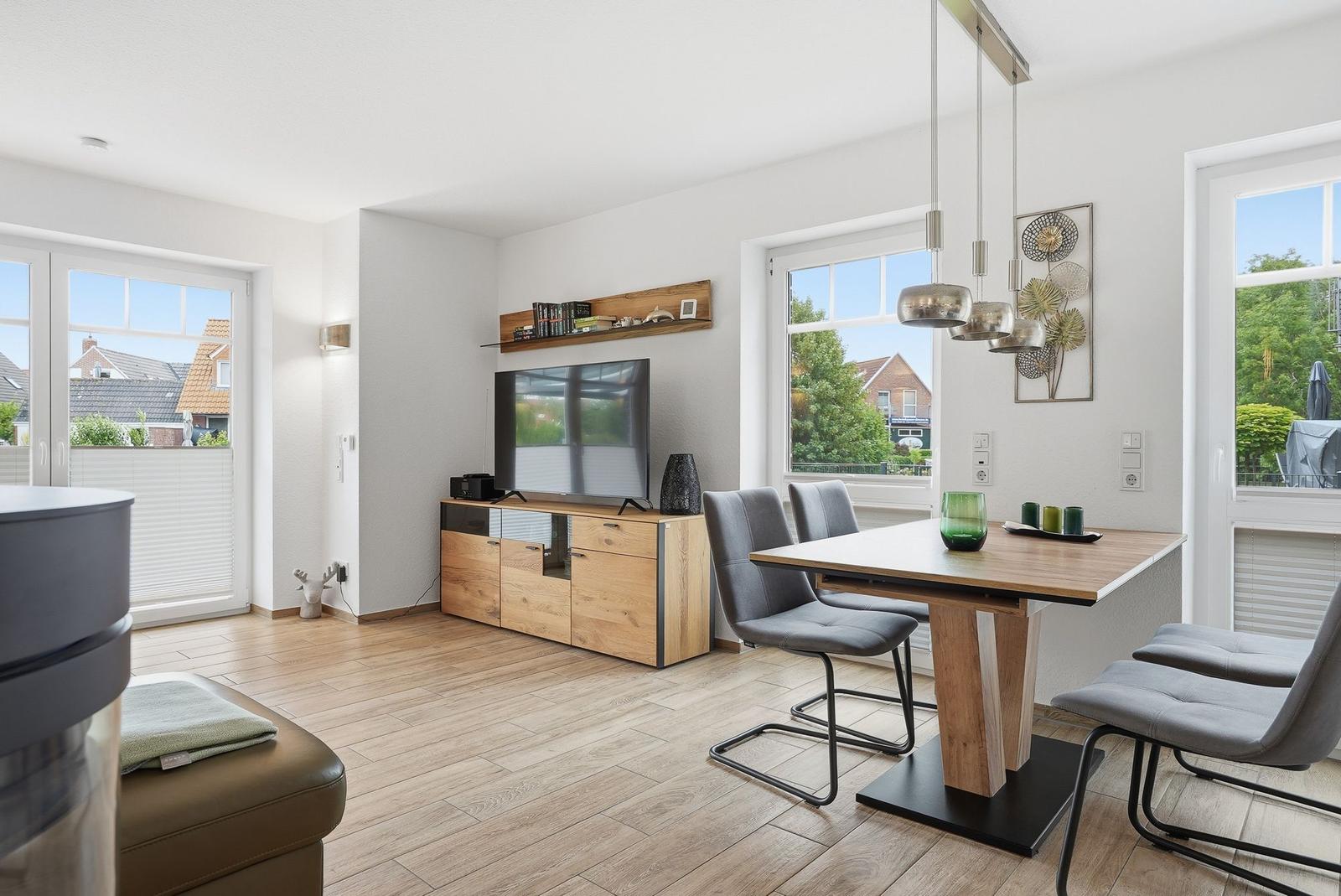 Wohn- und Essbereich mit TV, Tisch und Stühlen. Fenster bieten Blick auf Außenbereich.