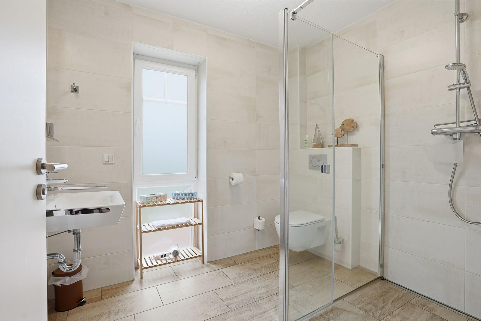 Badezimmer mit Dusche, WC und Waschbecken. Fenster, Holzregal und hell gefliest.